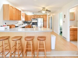 A modern kitchen with wooden cabinets, a central island with stools, a ceiling fan, and a view into the adjacent living area.