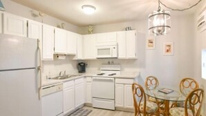 A compact kitchen with white cabinets, a microwave, a stove, and a dining area with a glass table and wicker chairs. - Ocean Block Condo, Steps from the beach, full kitchen, W/D, 1 Parking Spot , North Ocean City! (Ocean City)