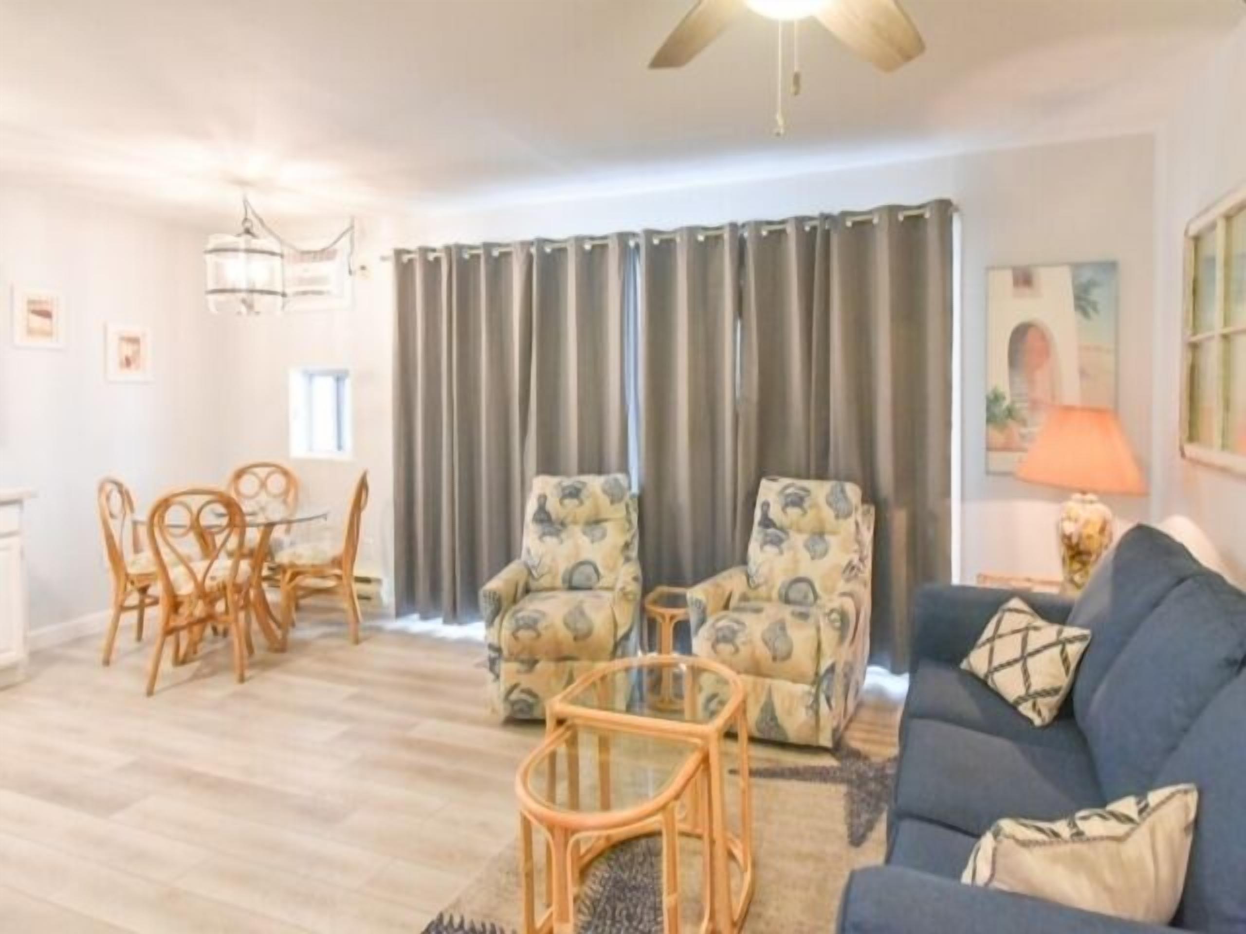 A living room with a blue sofa, patterned armchairs, a dining table with chairs, and a ceiling fan.