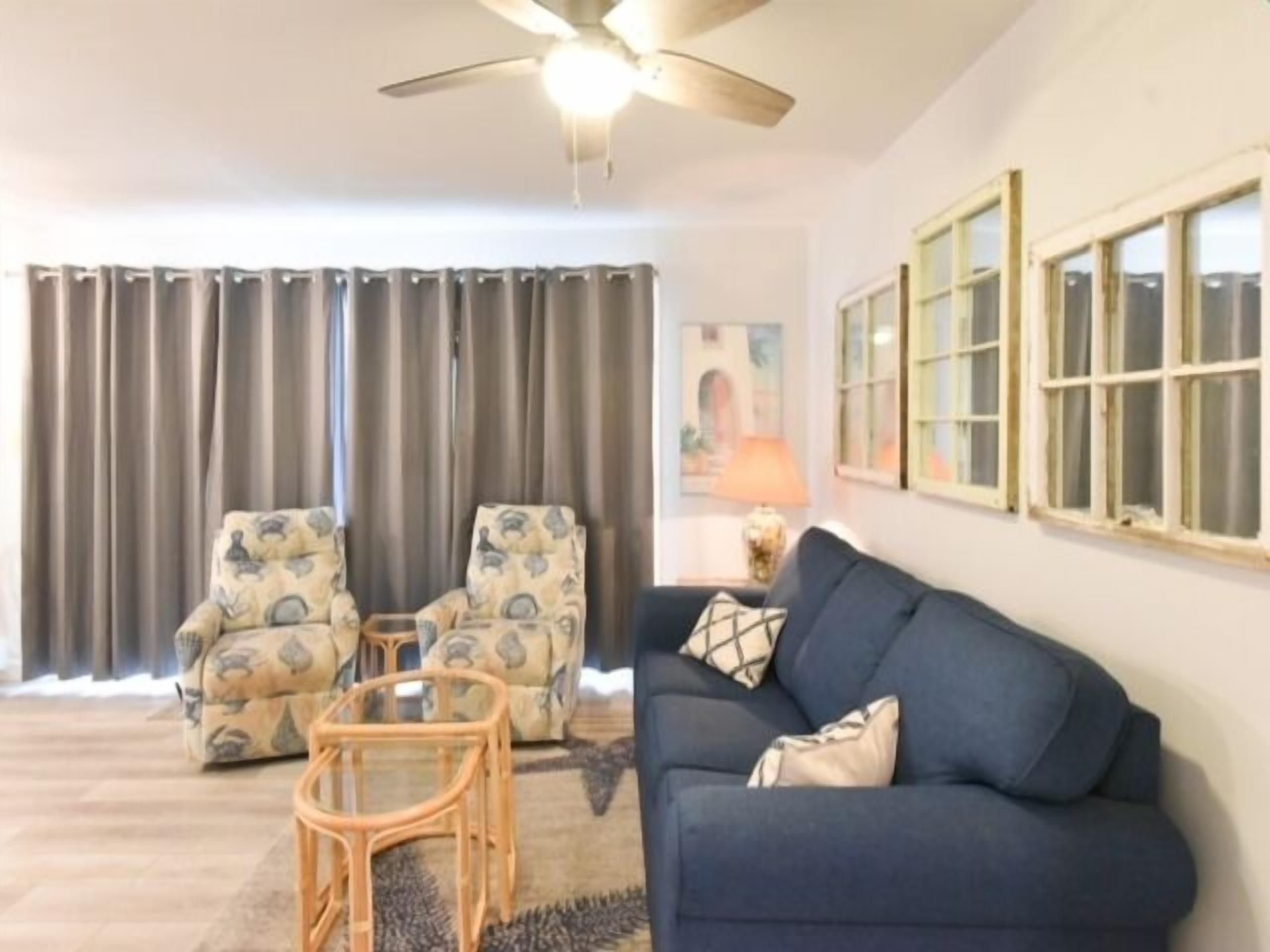Un salon avec un canapé bleu, deux fauteuils à motifs, une table basse en verre et un ventilateur de plafond.