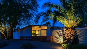 A modern house with a well-lit interior, surrounded by palm trees and a gravel driveway. - Stylish Mid Century Krisel Oasis -New pool deck, Salt Water Pool, Firepit, BBQ (Palm Springs)