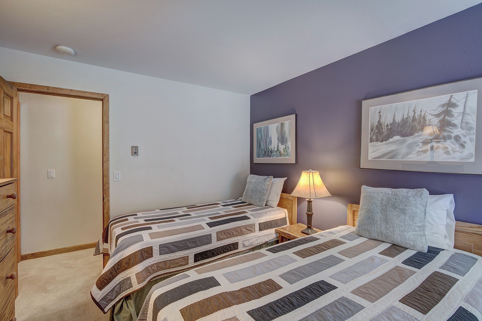 Une chambre avec deux lits, une commode en bois, une table de chevet avec une lampe, et deux tableaux encadrés accrochés au mur.