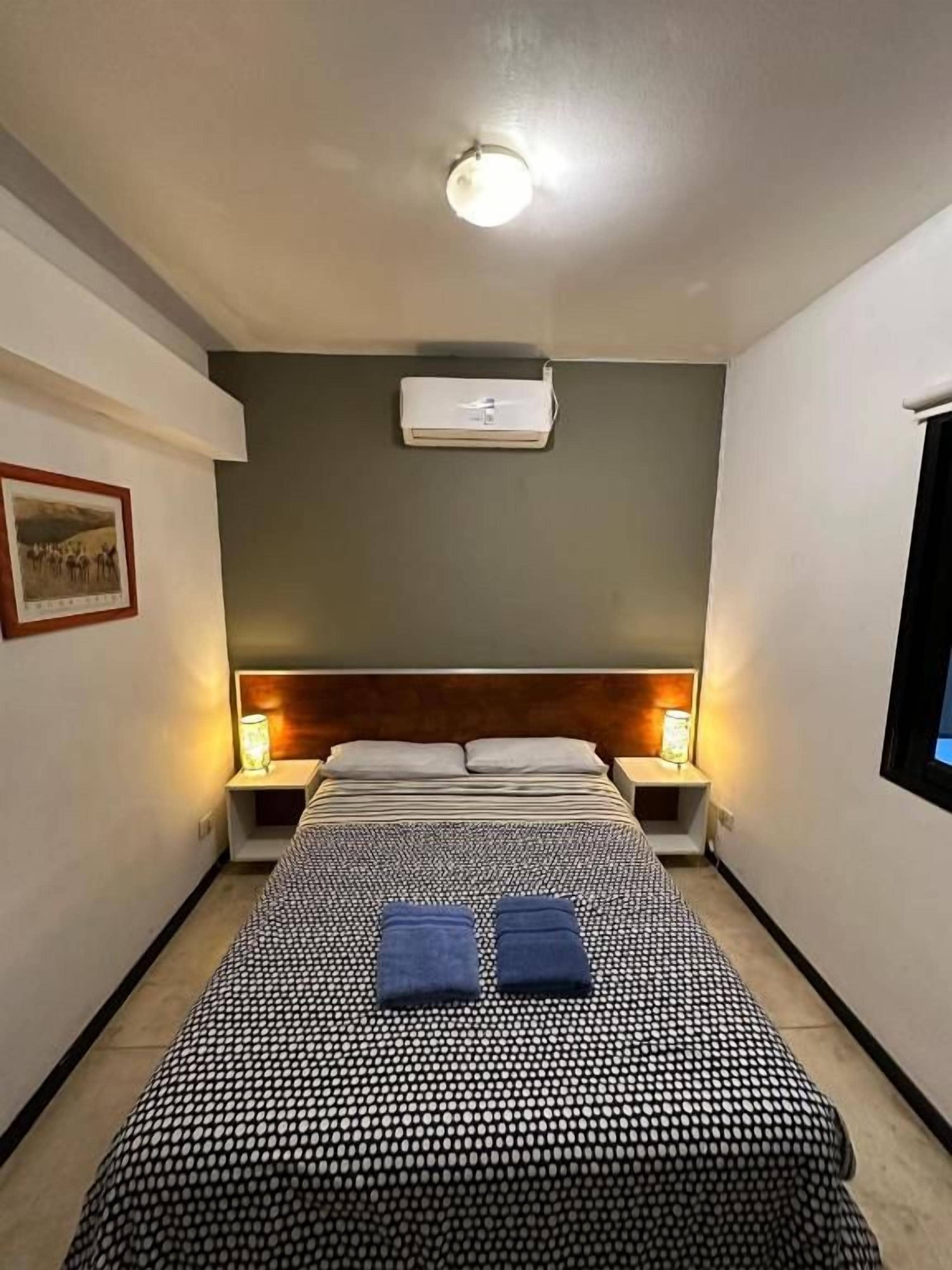 A hotel room with a bed, two bedside lamps, an air conditioner, and a framed picture on the wall.