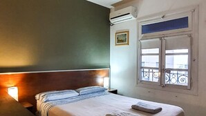 A bedroom with a bed, bedside table, ceiling fan, air conditioning unit, window with bars, and a picture on the wall. - Hostel Carlos Gardel (Buenos Aires)