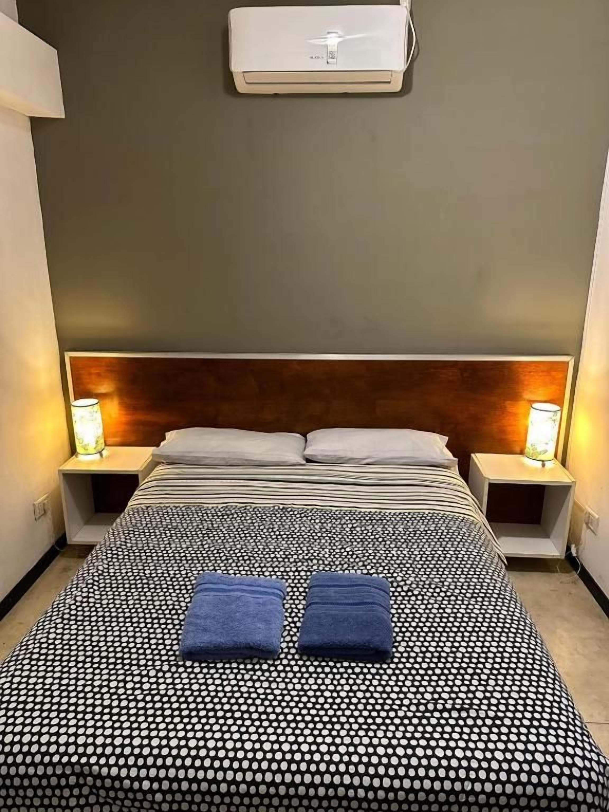 A hotel room with a bed, two bedside lamps, and an air conditioning unit.