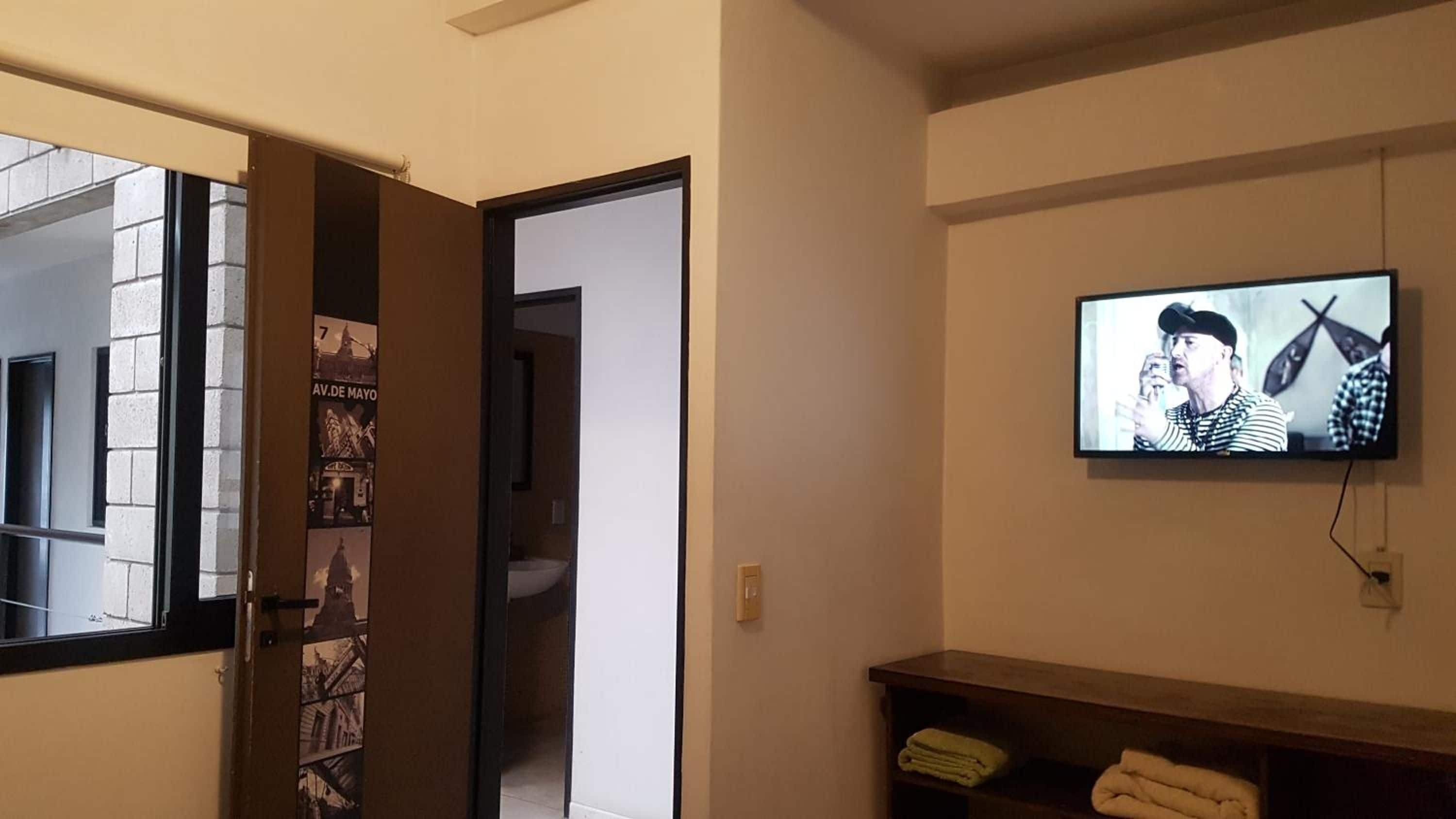 Une chambre d’hôtel avec un téléviseur fixé au mur, une porte donnant sur une autre pièce et une étagère où sont pliées des serviettes.