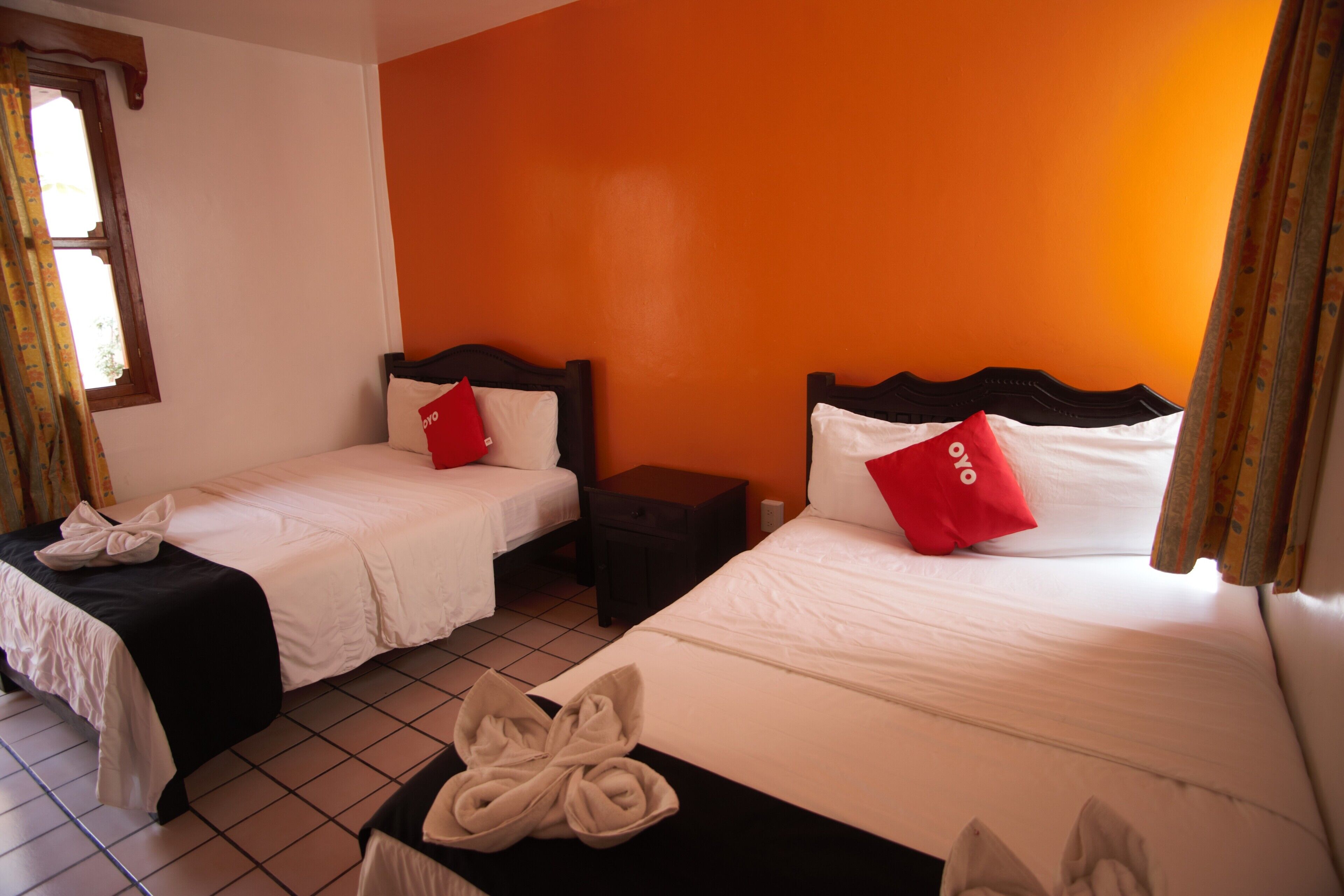 Hotelkamer met twee bedden, elk met wit beddengoed en rode kussens, een klein houten nachtkastje en een raam met gordijnen.