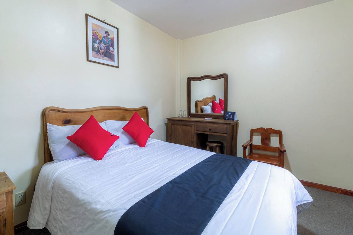 A neatly made bed with white and navy bedding, a wooden headboard, and two red pillows. A wooden nightstand with a lamp is to the left. A wooden dresser with a mirror is against the wall.