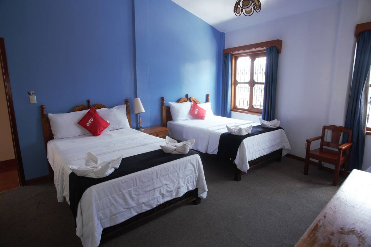 A hotel room with two single beds, a wooden chair, and a window with blue curtains.
