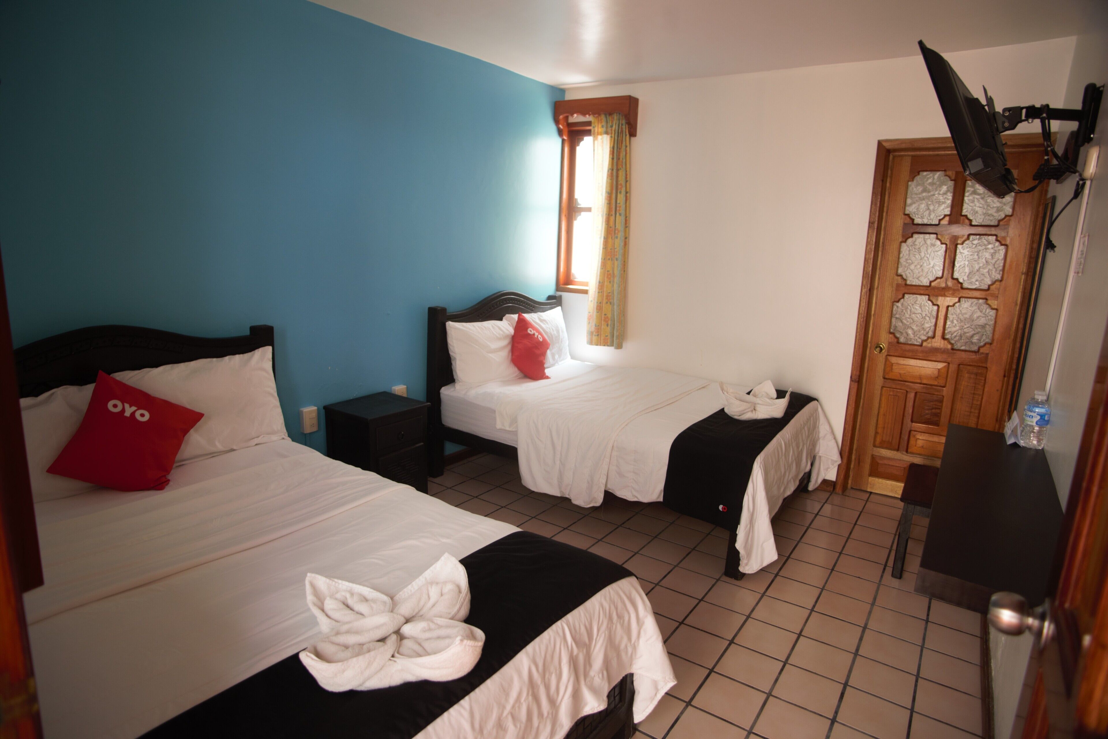 A room with two beds, a wooden door, a window with curtains, and a ceiling-mounted light fixture.
