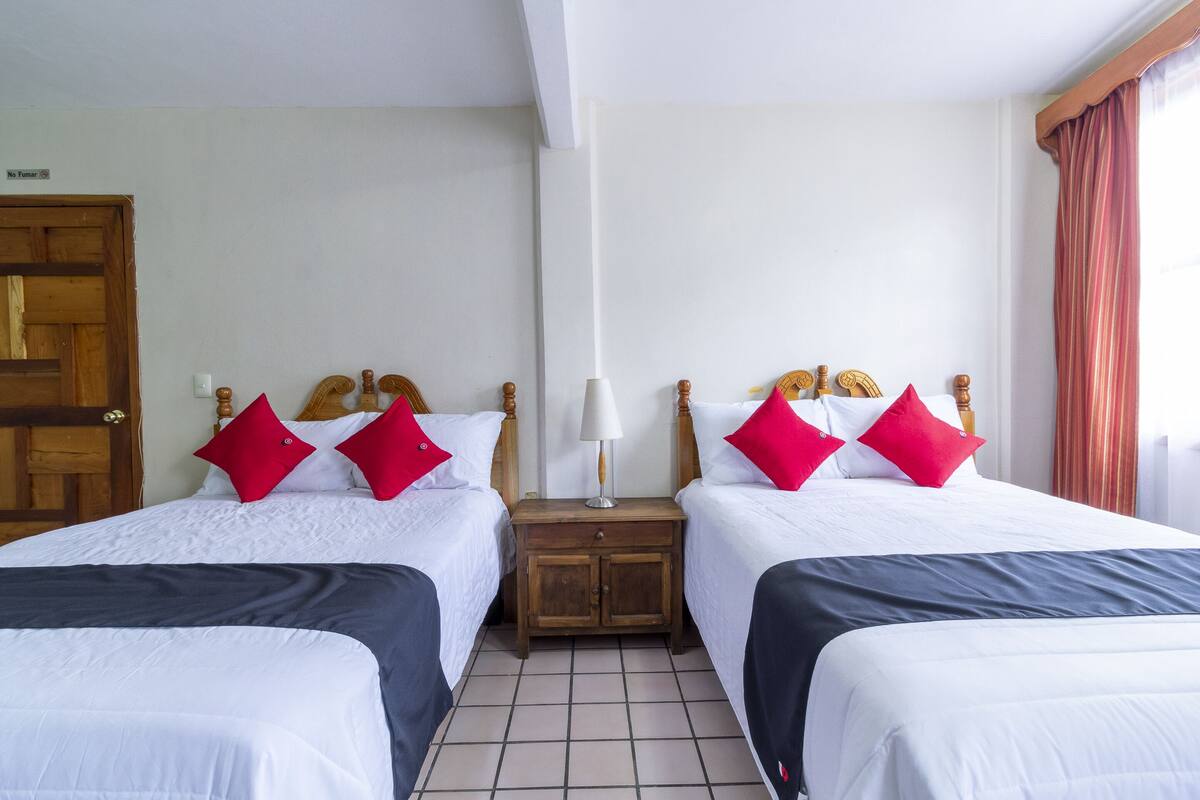 A hotel room with two beds, each with white bedding and red pillows, a wooden nightstand with a lamp, and a tiled floor.
