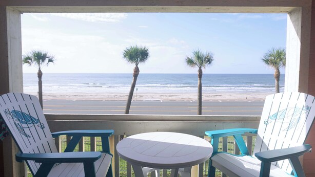 Un balcon agrémenté de deux fauteuils Adirondack et d’une petite table ronde, offrant une vue sur une plage bordée de palmiers et sur l’océan.