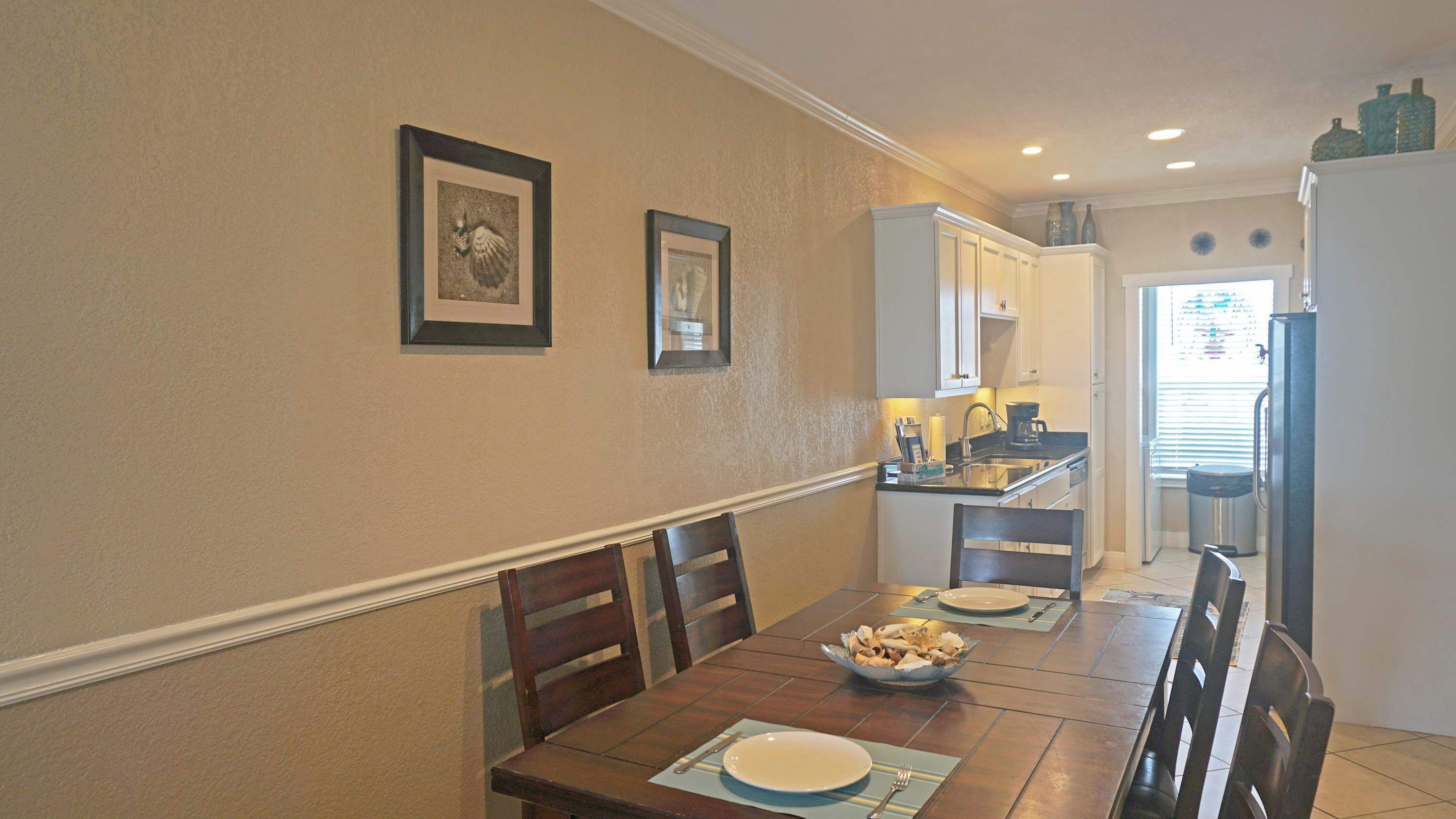 A dining area with a wooden table set for two, framed pictures on the wall, and a kitchen in the background.