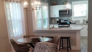 A spacious living area with a patterned sofa, a round coffee table, and a dining table with chairs. There is a kitchen area with a microwave and a refrigerator in the background.