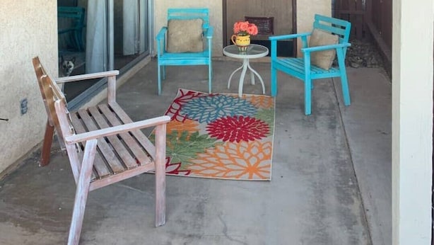 Un espace extérieur abrité, avec un banc en bois, deux chaises bleues, une petite table ronde ornée d’un vase de fleurs et un tapis coloré.