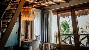 A room with a wooden ceiling structure, a concrete bench, a wicker basket, and a view of palm trees through large windows. - Aldea Kuká, Luxury Eco Boutique Hotel  (Isla Holbox)