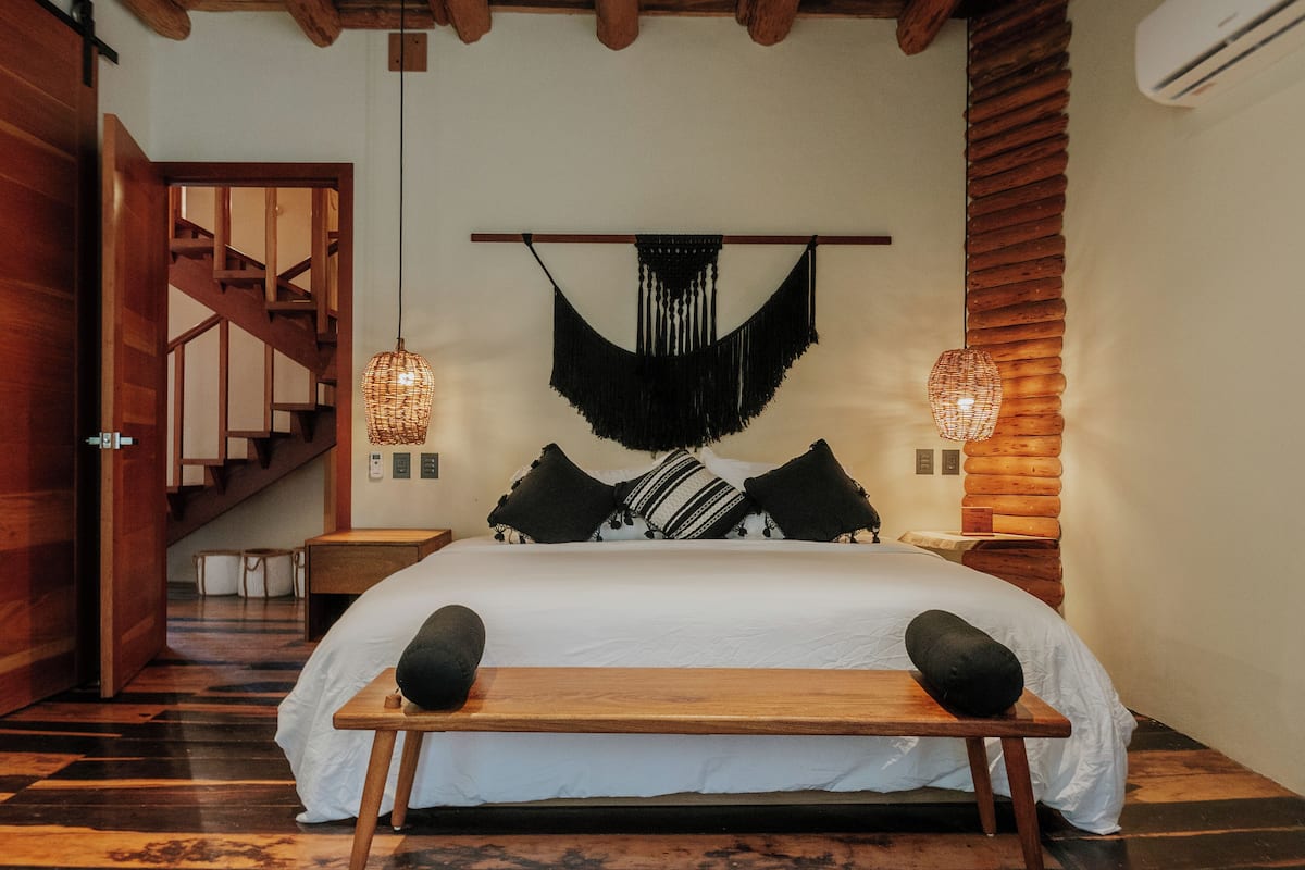 A bedroom with a wooden bed, a bench, a hanging macrame wall decoration, and a staircase.