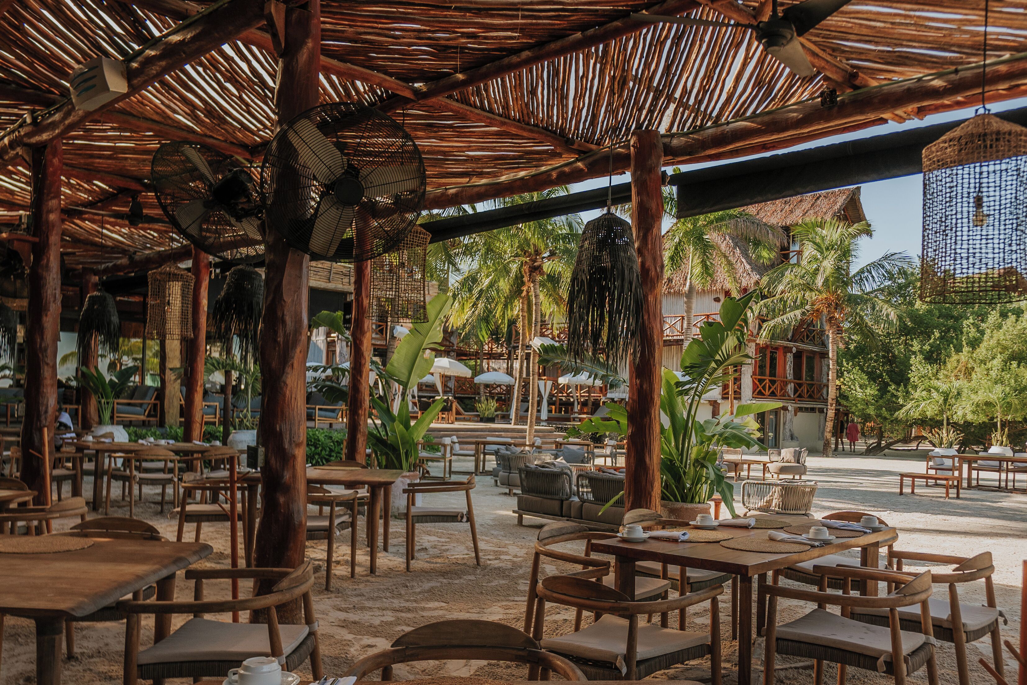 An outdoor dining area with wooden tables and chairs, surrounded by palm trees and a thatched roof structure.