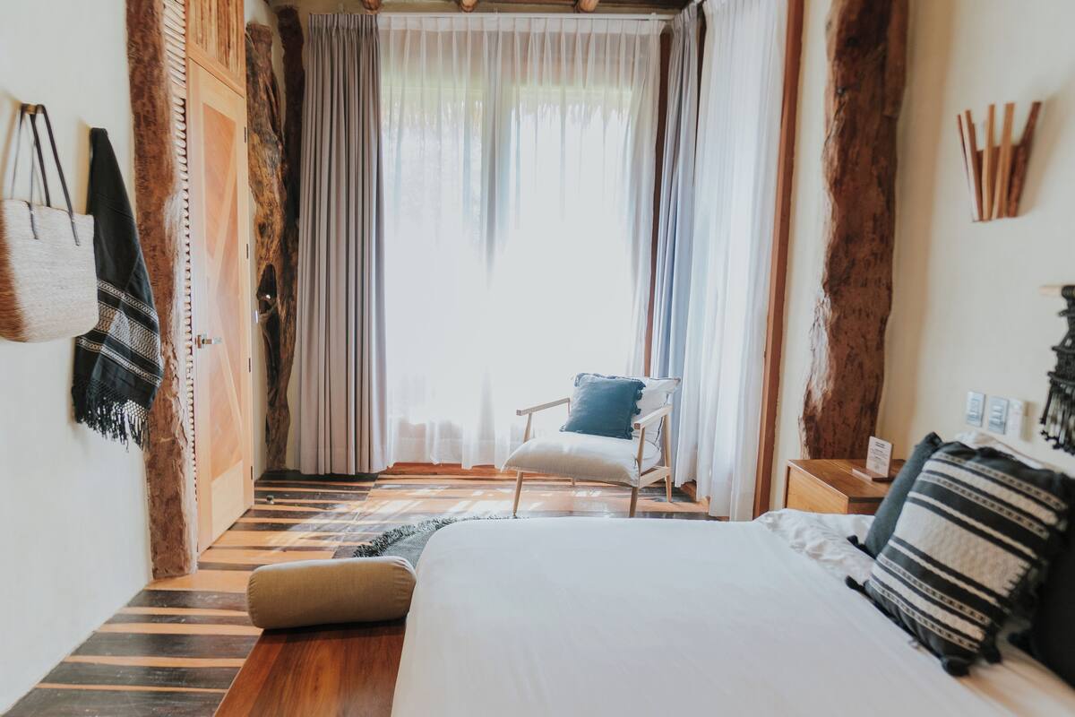 A bedroom with a large bed, a chair, a nightstand, and a window with curtains.