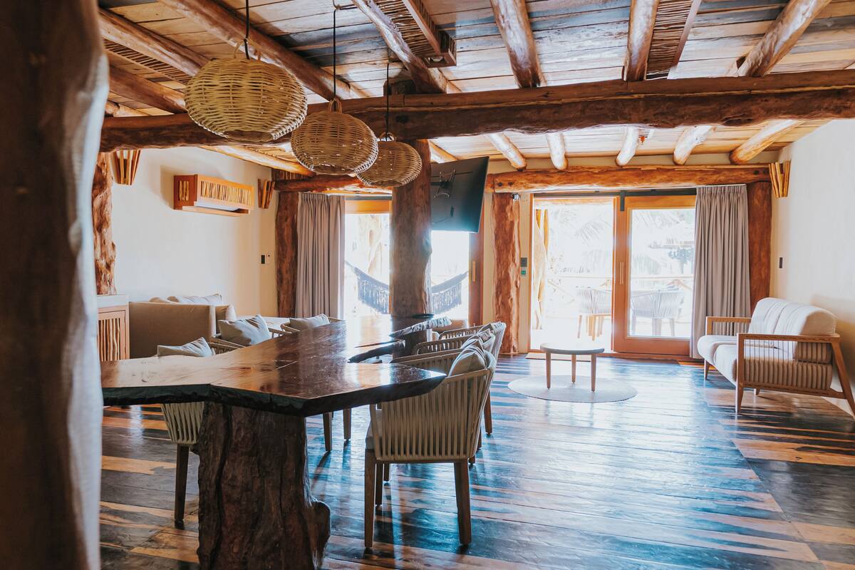 A spacious room with a large wooden dining table, wicker hanging lights, and a view through a glass door.