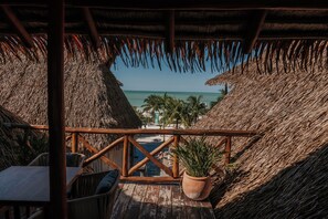 A wooden deck with a thatched roof, overlooking a beach and palm trees.