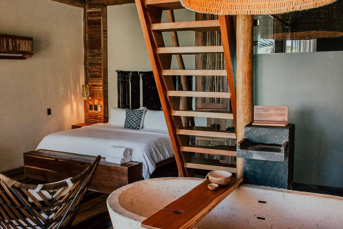 A room with a bed, a wooden chair, a woven light fixture, a wooden ladder, and a small table with a bowl.