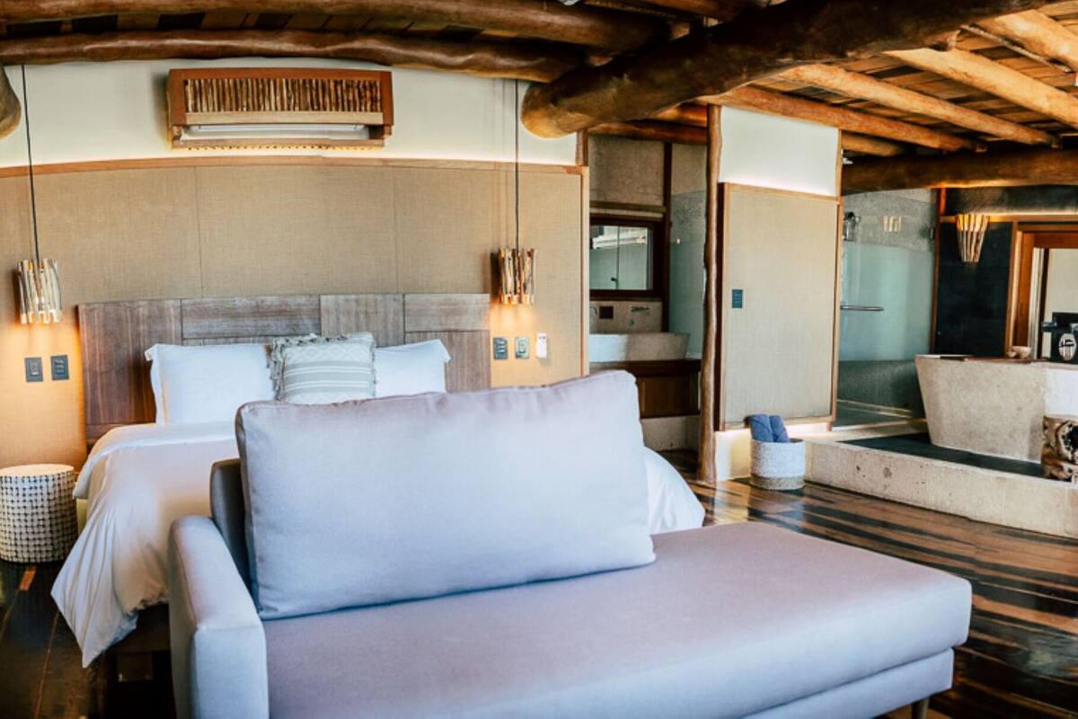 A modern hotel room with a large bed, a sofa, a wooden ceiling, and a view of the outdoors.
