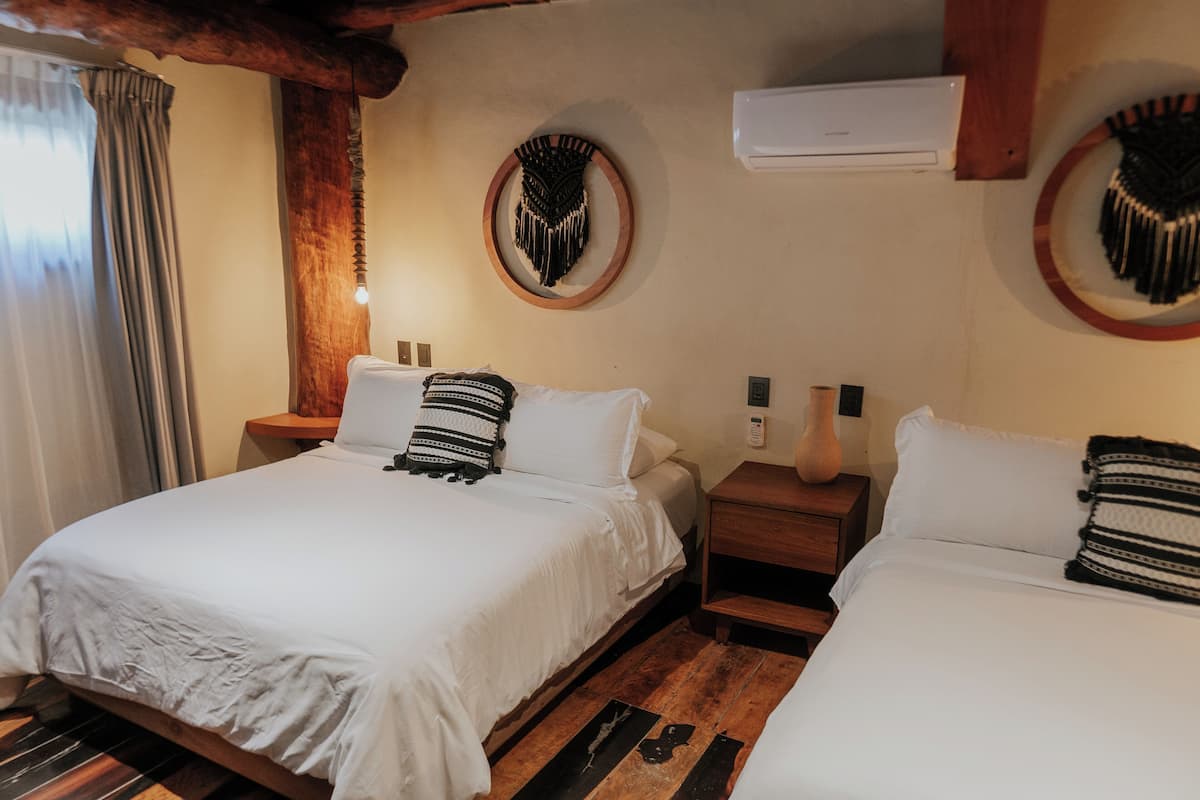 A room with two beds, wooden flooring, a wooden ceiling, a wall-mounted air conditioner, and a window with curtains.