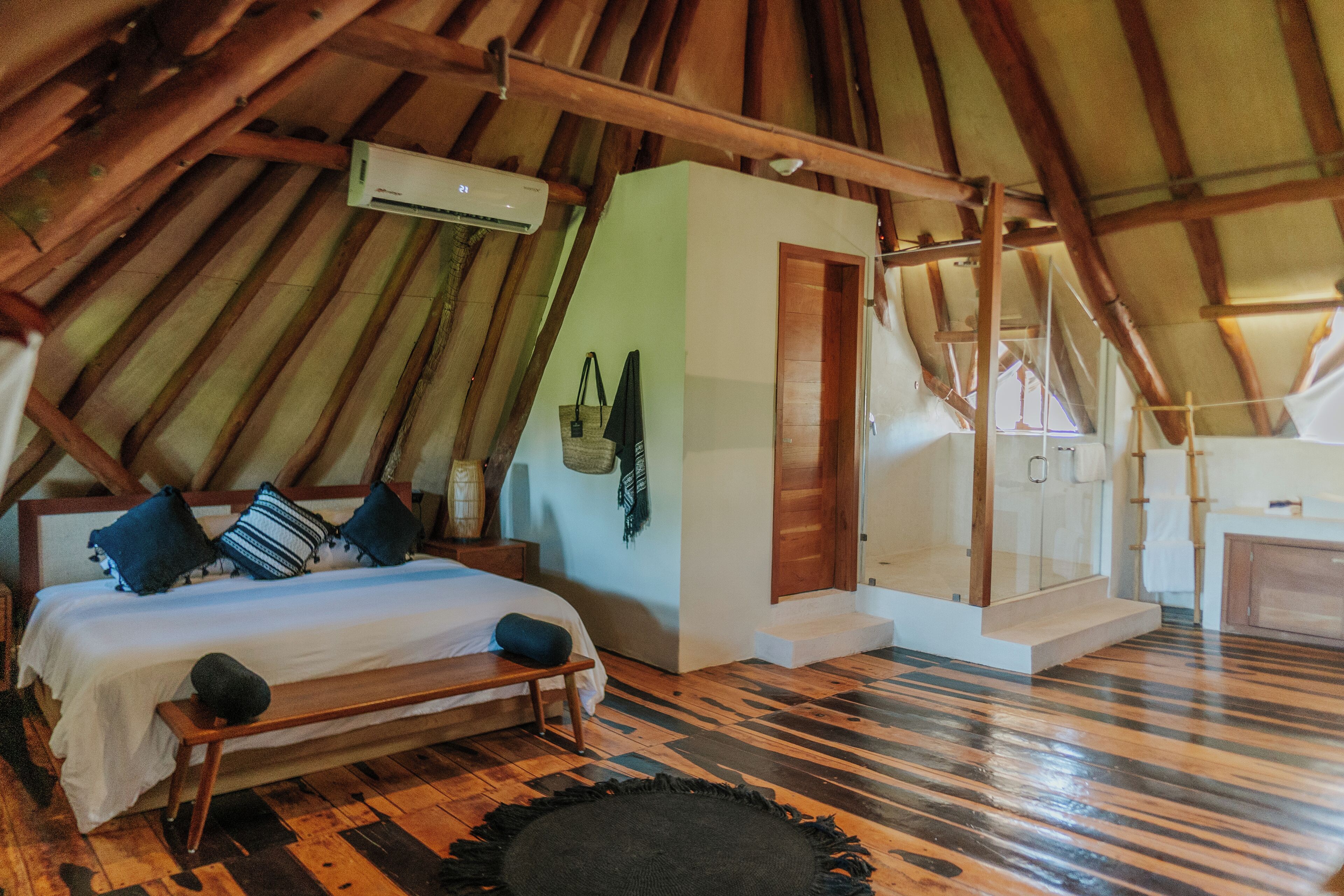 A bedroom with a wooden ceiling, a bed with pillows, a bench, a small table, and a wall-mounted air conditioner.