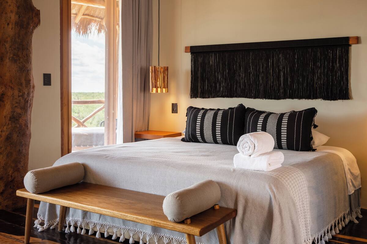 A bedroom with a large bed, a bench, a view of the outdoors, and a wooden headboard.