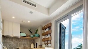 A modern kitchen with white cabinets, a wooden stool, and a view of the ocean through a sliding glass door. - The Beach House - Studios & Suites (Javea)
