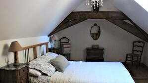 A bedroom with a sloped ceiling, a bed with a floral-patterned bedspread, a nightstand with a lamp, a wooden chair, and a small table with a mirror. - Luxury gites & B&B, near Saumur, in 18thC former wine estate, heated pool  (Les Bois D'anjou, Pays De La Loire)