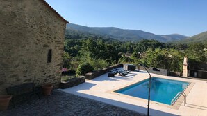 Un bâtiment en pierre doté d’une piscine, de chaises longues et offrant une vue sur les montagnes et les éoliennes.