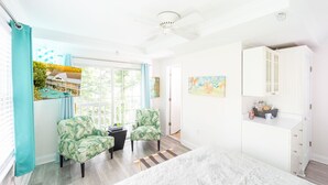 Ein helles Zimmer mit einem Bett, zwei Stühlen, einem kleinen Tisch, einem Gemälde und Blick auf ein Haus durch das Fenster.