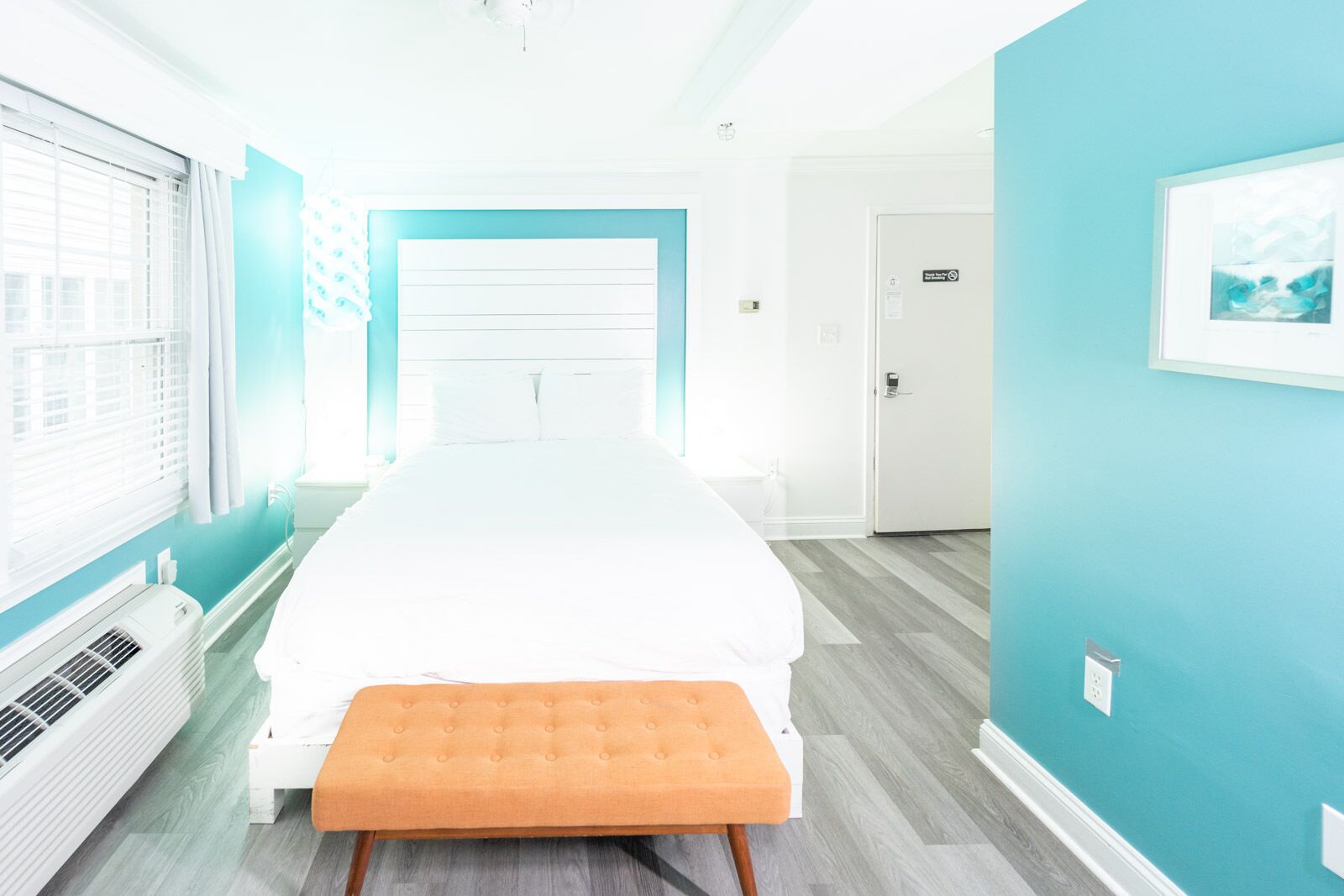 A hotel room with a large bed, a bench, a window with blinds, and a door.
