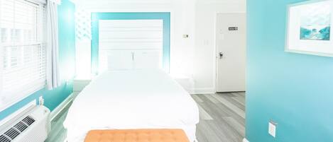 A hotel room with a large bed, a bench, a window with blinds, and a door.
