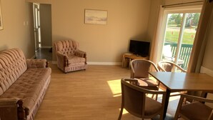 A living room with a sofa, armchair, dining table, and chairs. There is a television on a stand and a painting on the wall. - Gateway Motel (Minnedosa)