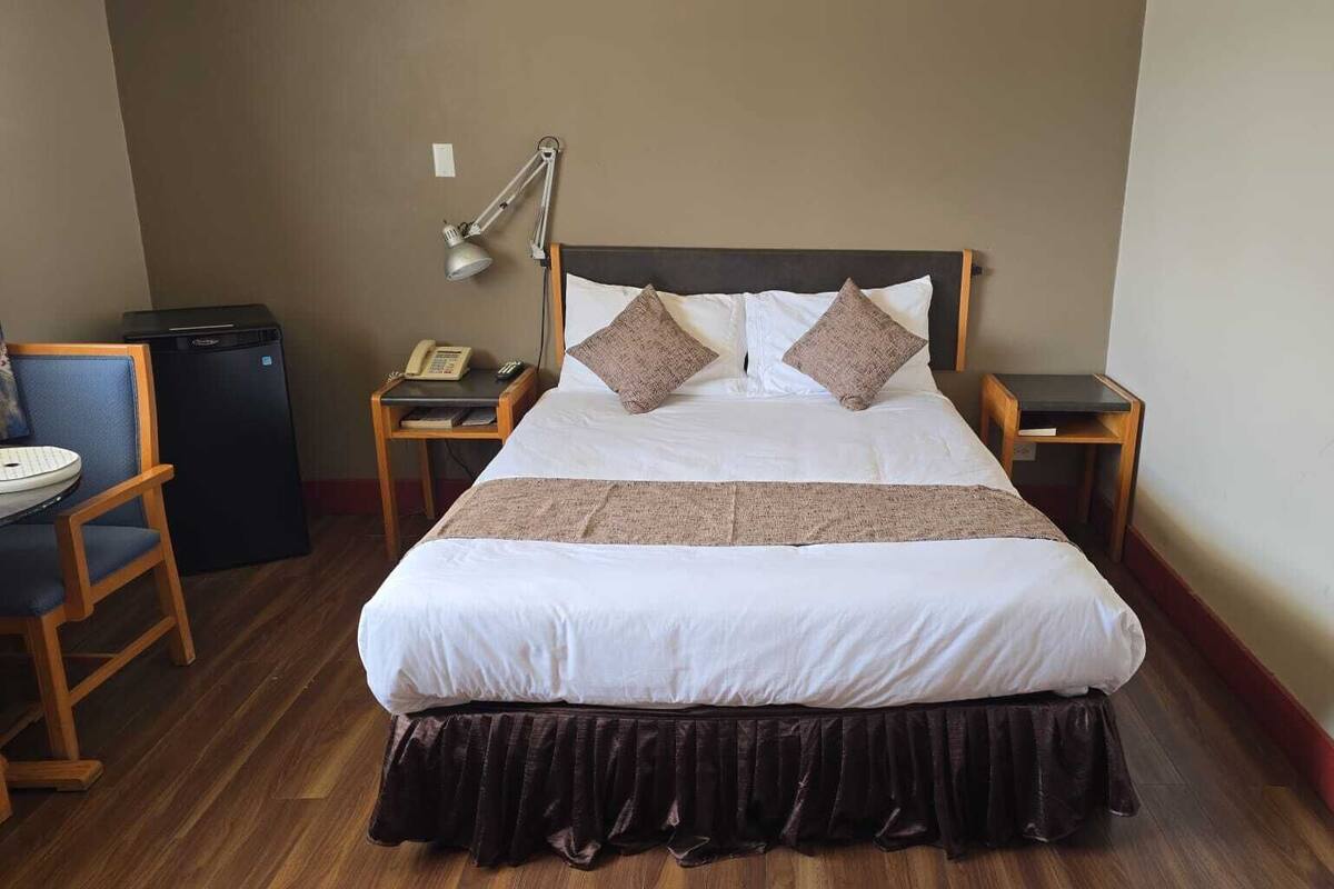 A neatly made bed with a headboard, two bedside tables, a desk, a chair, and a small refrigerator.