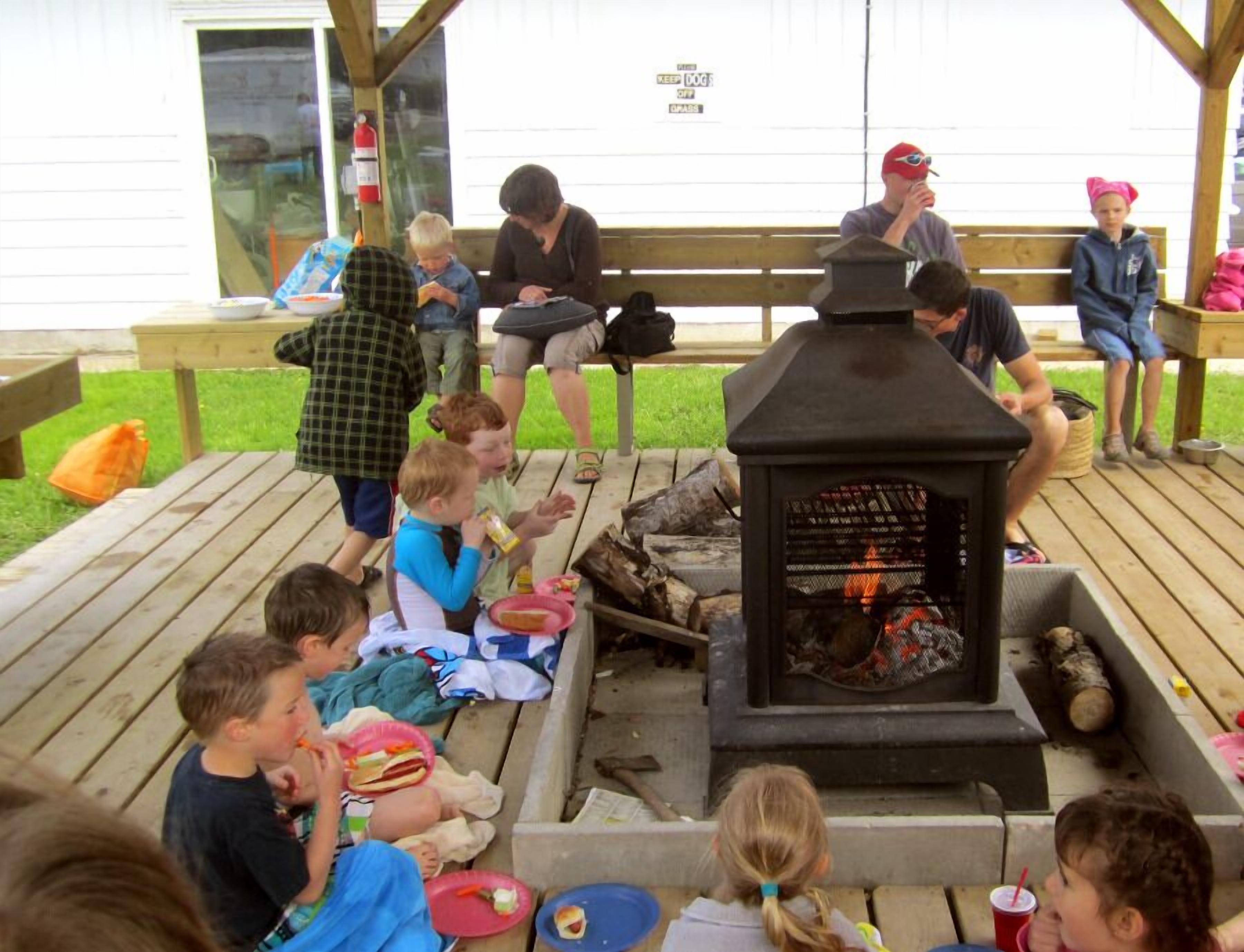 A group of people sitting around a wood-burning stove on a wooden deck, with a sign that reads "KEEP DOG OFF GRASS".