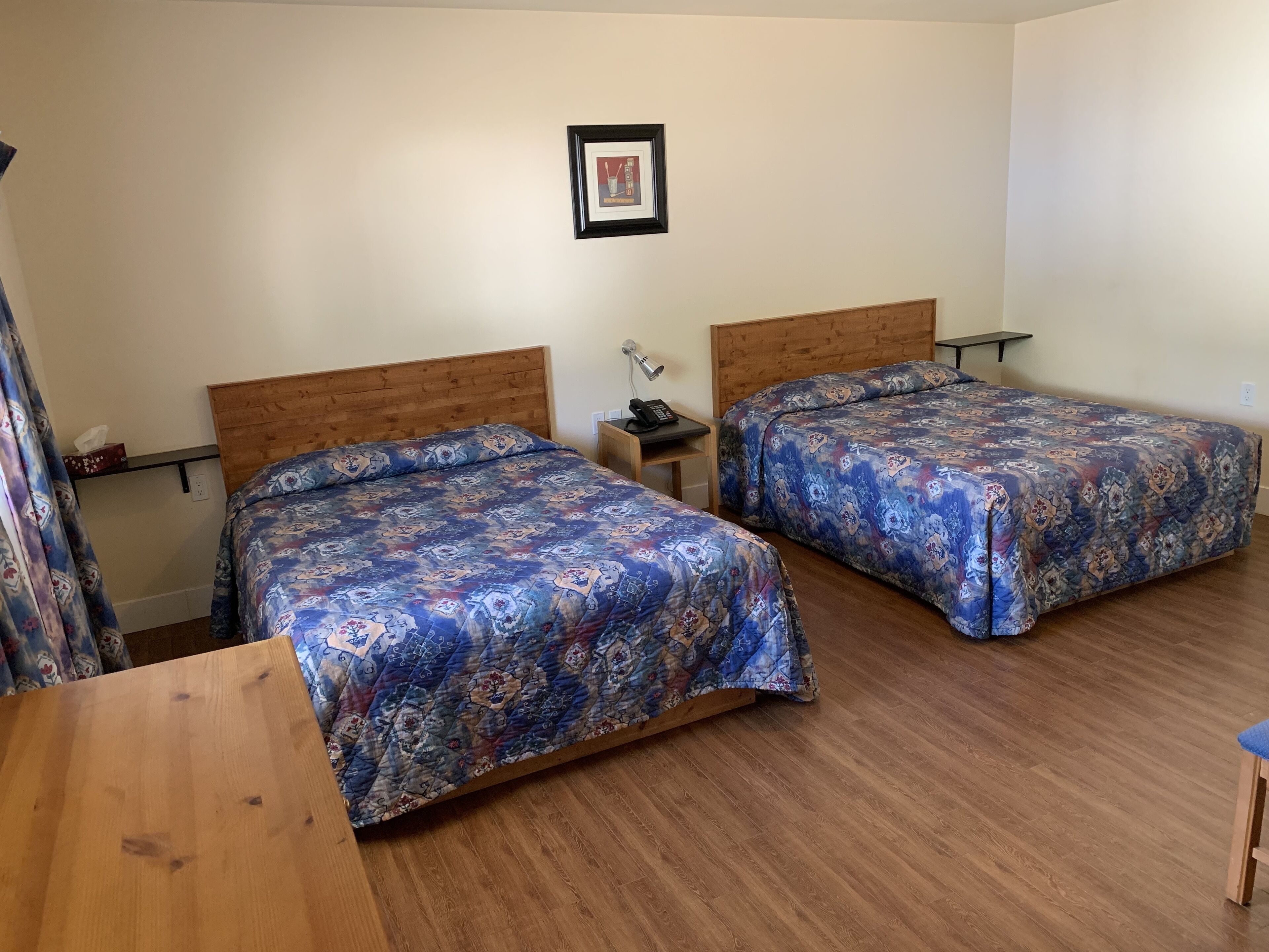 Hotellihuone, jossa on kaksi sänkyä, puiset päätyosat, yöpöytä lampun kanssa ja seinällä kehystetty kuva.