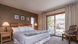 Ein Schlafzimmer mit einem großen Bett, zwei Nachttischen mit Lampen, einem Sessel und Blick auf eine gebirgige Landschaft durch große Fenster.