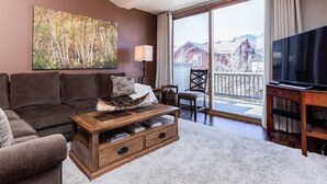 Ein Wohnzimmer mit einer Couch, einem Couchtisch und einem Gemälde an der Wand. Ein Fernseher auf einem Schrank und ein Fenster mit Blick auf Gebäude.
