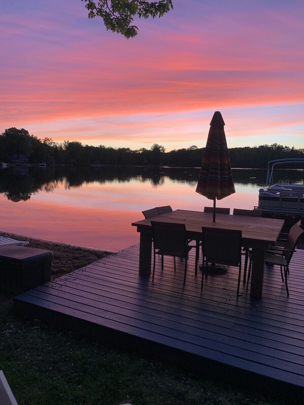 A wooden deck with a table and chairs, an umbrella, and a boat docked by the water. - Best Lake in Southwest Michigan! Pontoon Boat included. Private beach frontage. (Grand Junction)