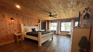 A wooden-paneled room with a bed, a small table, a nightstand, a ceiling fan, and a window with curtains. - Best Lake in Southwest Michigan! Pontoon Boat included. Private beach frontage. (Grand Junction)