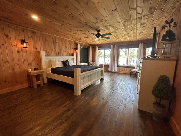 A wooden-paneled room with a bed, a small table, a nightstand, a ceiling fan, and a window with curtains. - Best Lake in Southwest Michigan! Pontoon Boat included. Private beach frontage. (Grand Junction)