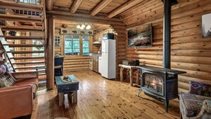 Een houten blokhut van binnen met een houten vloer, een open haard, een bank, een eettafel en een keukenhoek.