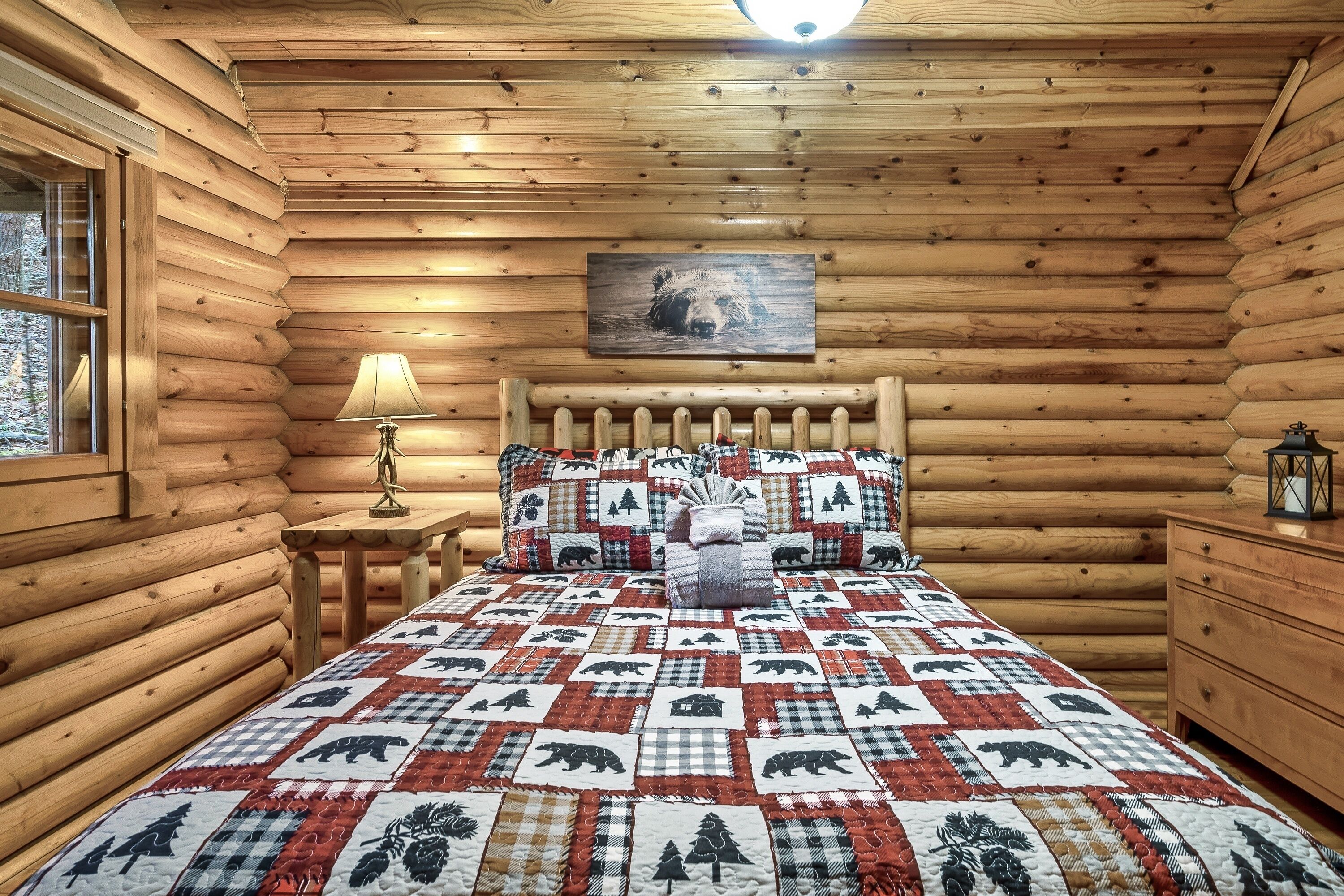 A log cabin bedroom with a quilted bedspread featuring bear and tree patterns, a wooden headboard, a bedside lamp, and a framed bear picture on the wall.