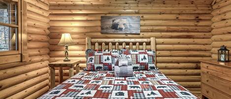 A log cabin bedroom with a quilted bedspread featuring bear and tree patterns, a wooden headboard, a bedside lamp, and a framed bear picture on the wall.