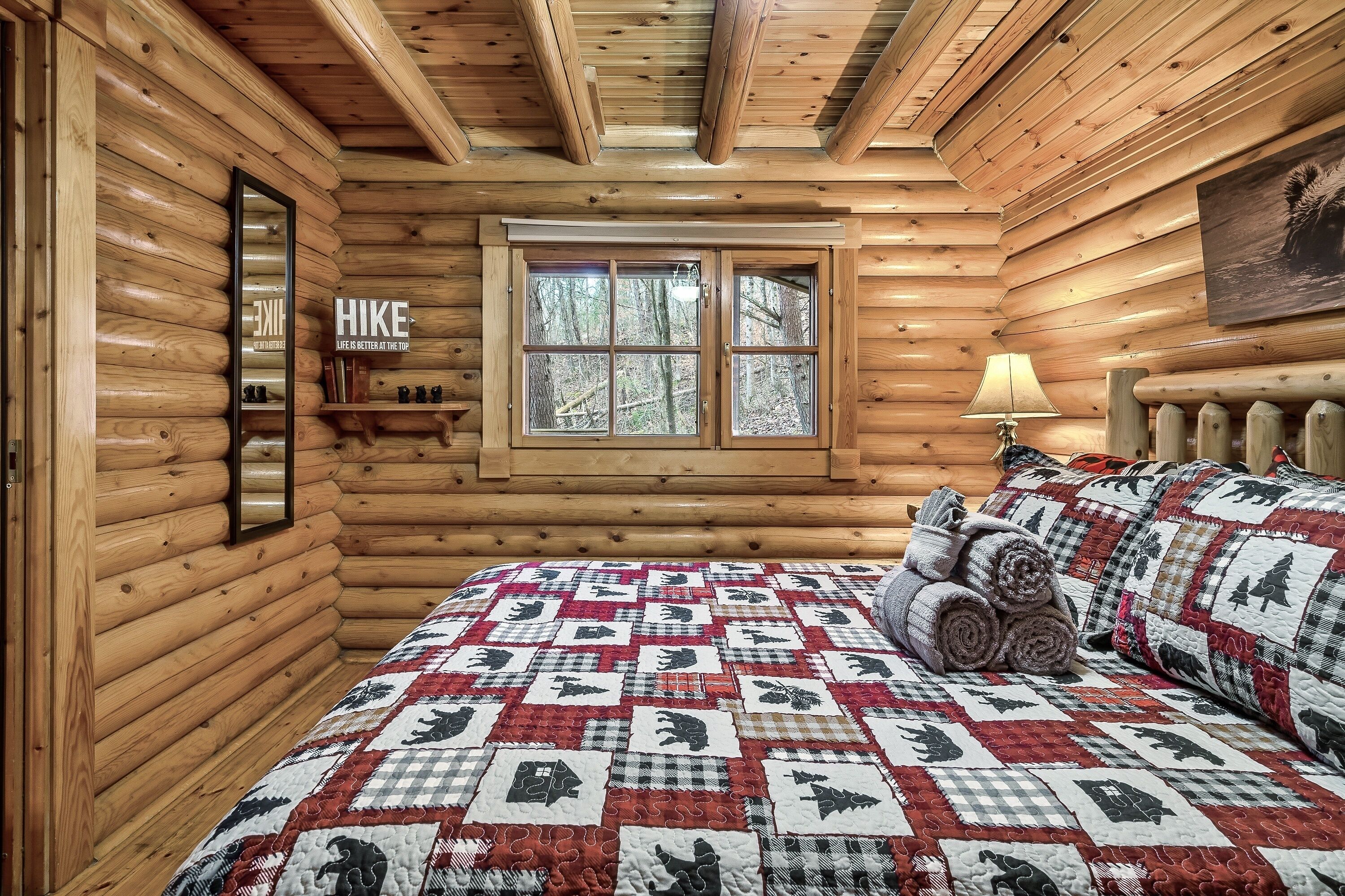 A log cabin bedroom with a quilted bedspread, a window with a view of trees, and a mirror on the wall.