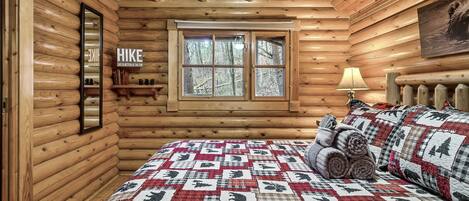 A log cabin bedroom with a quilted bedspread, a window with a view of trees, and a mirror on the wall.